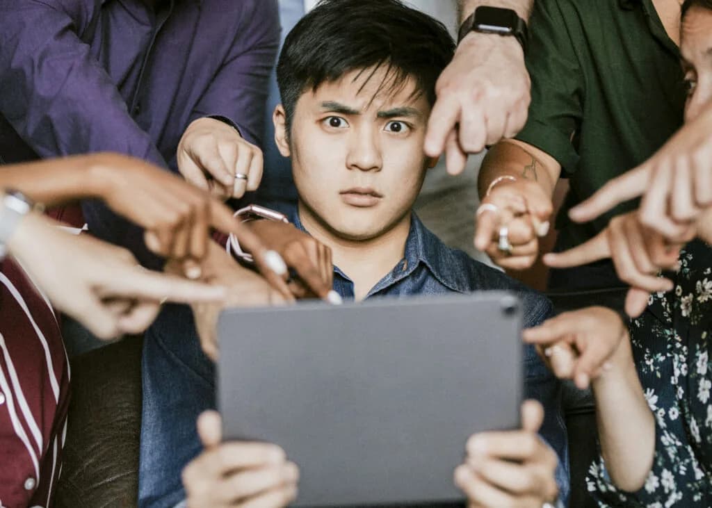 Un homme assis tient une tablette et regarde l’écran avec un air choqué. Autour de lui, plusieurs personnes tendent leurs bras et pointent du doigt vers la tablette, comme si elles lui mettaient la pression ou lui demandaient quelque chose.