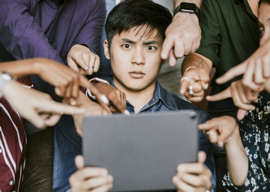 Un homme assis tient une tablette et regarde l’écran avec un air choqué. Autour de lui, plusieurs personnes tendent leurs bras et pointent du doigt vers la tablette, comme si elles lui mettaient la pression ou lui demandaient quelque chose.